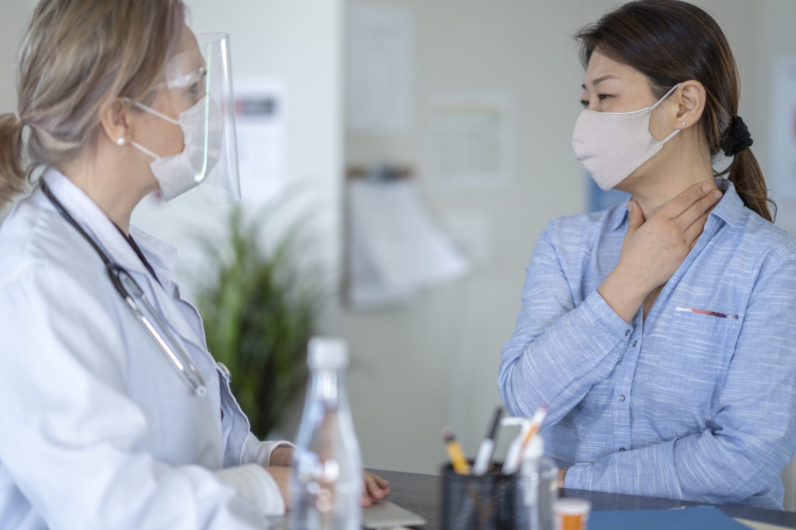 A female adult of Asian descent is at a medical check up at a doctor's office. She has one hand on her neck as she explains her medical condition. Her doctor is listening intently to her patient.