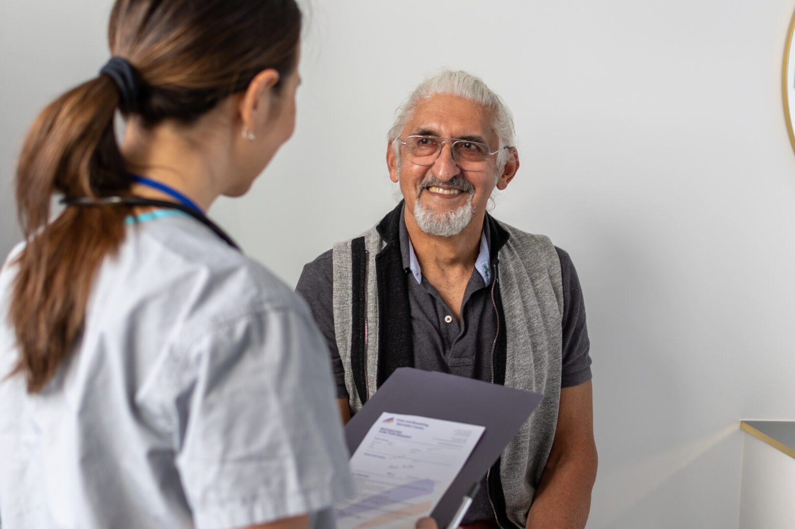 Focus on a senior male of Indian ethnicity with grey hair. He is smiling at the healthcare professional who is collecting his personal information. Medical clinic setting.