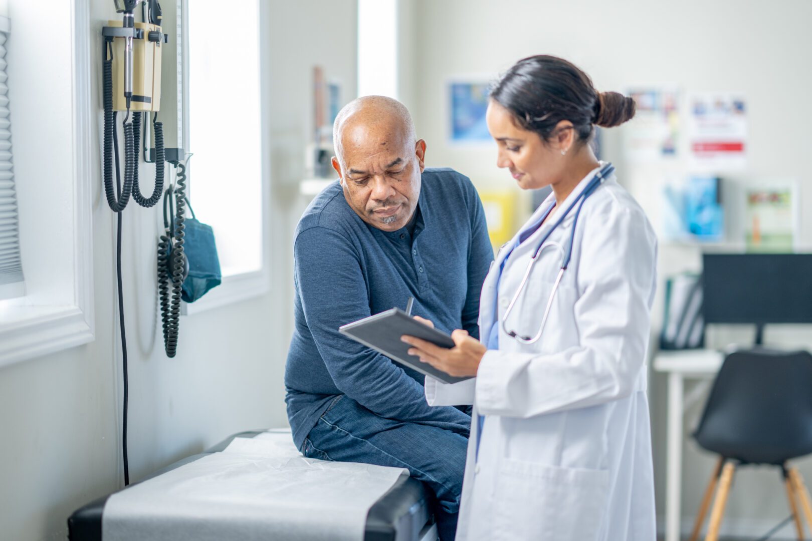 A senior gentleman of African decent sits up on an exam table during a routine check-up.  His female doctor is holding out a tablet as they review some recent test results together.