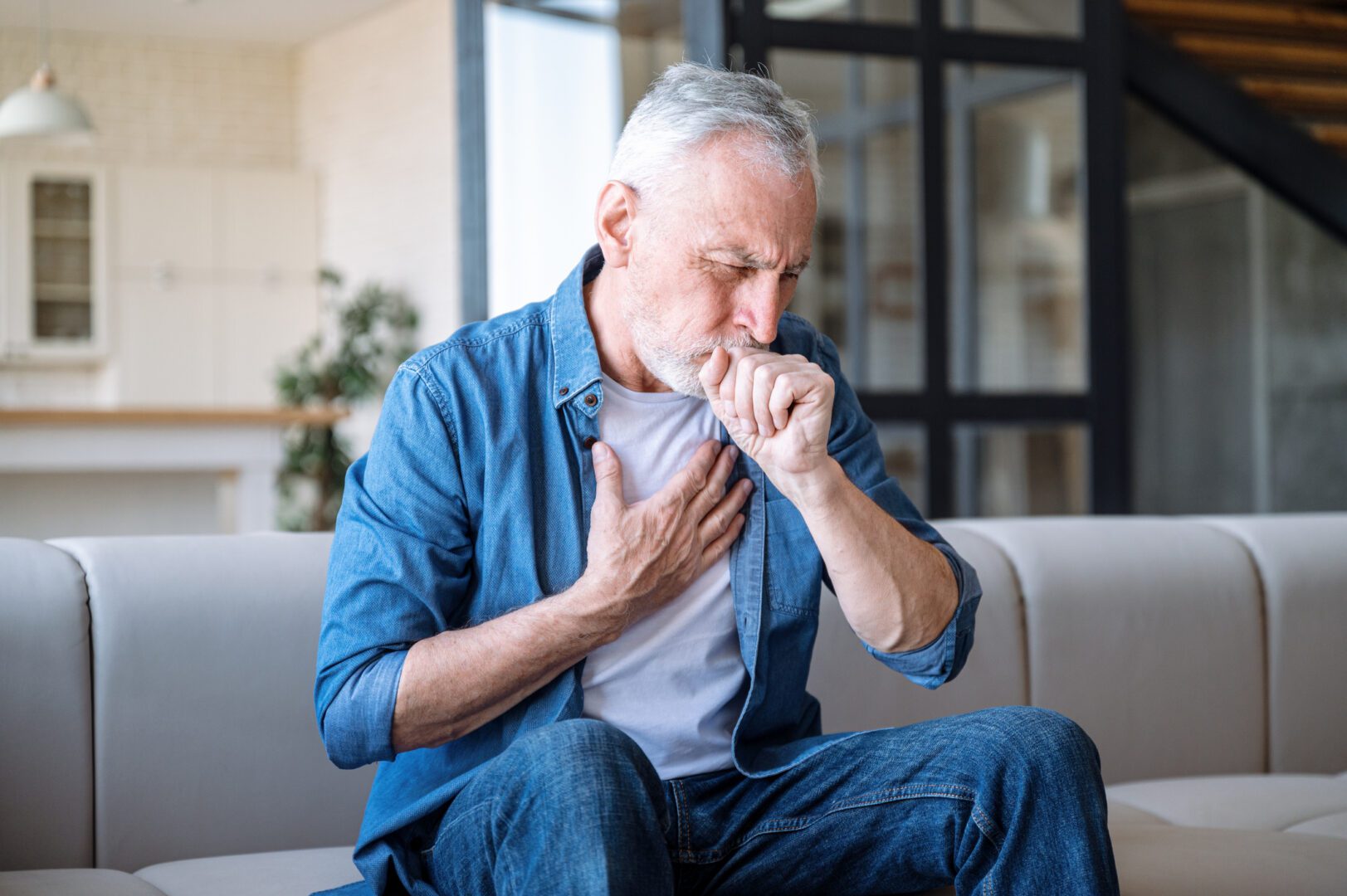 Senior man sitting on sofa at home and holding hand on chest. Male having asthma attack, difficulties with breathe, feeling severe pain or dyspnea