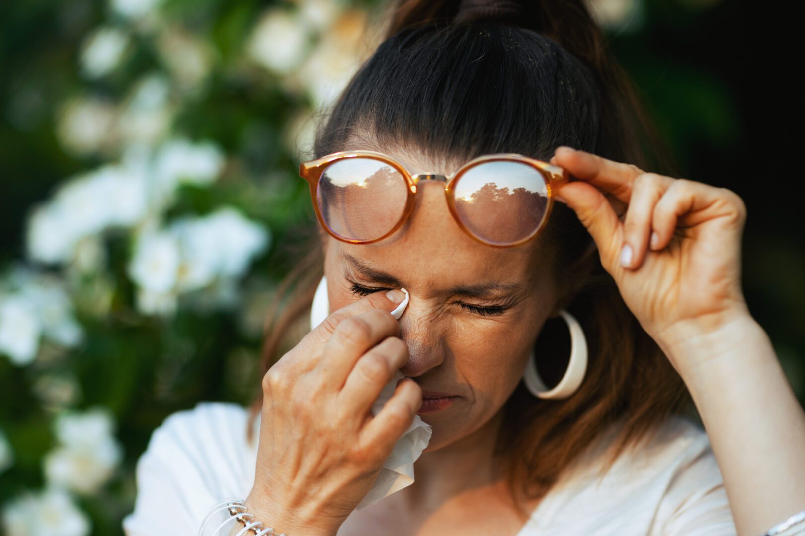 Summer time. sad modern 40 years old woman in white shirt with handkerchief and eyeglasses has an allergy attack near flowering tree.