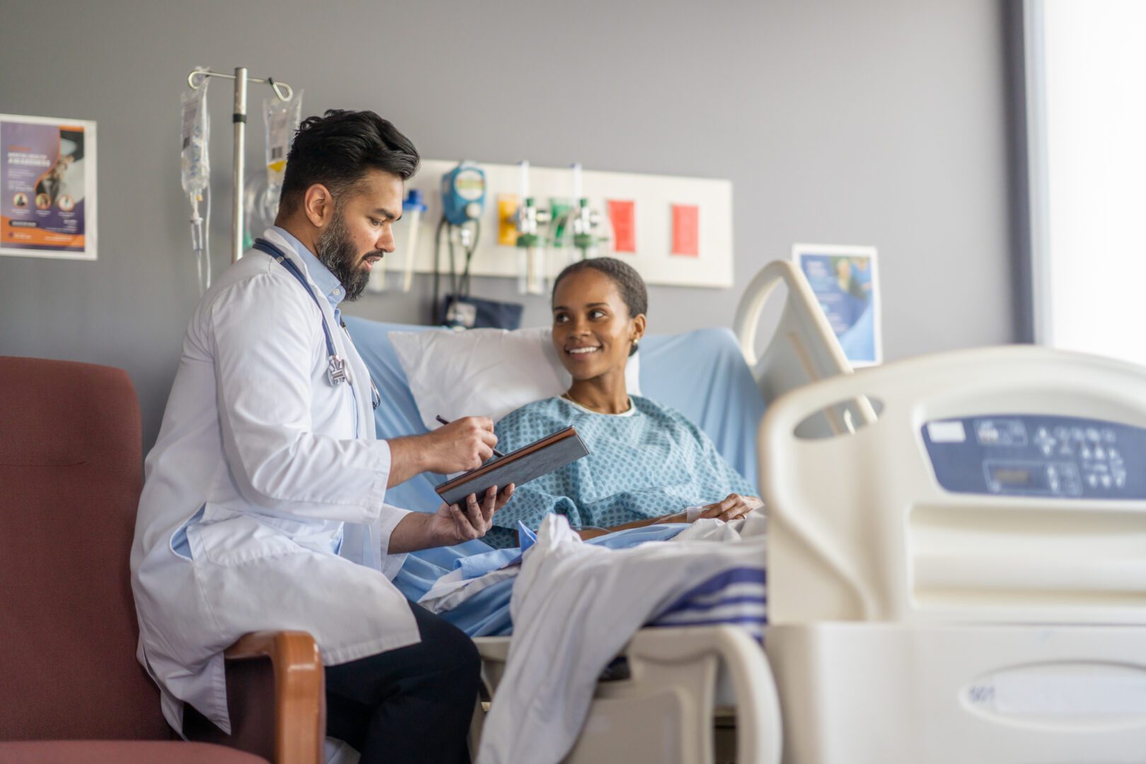 A woman sits up in a hospital bed wearing a gown as she talks with her male doctor about her surgery.  The doctor is taking notes electronically on a tablet as the two talk about the procedure.