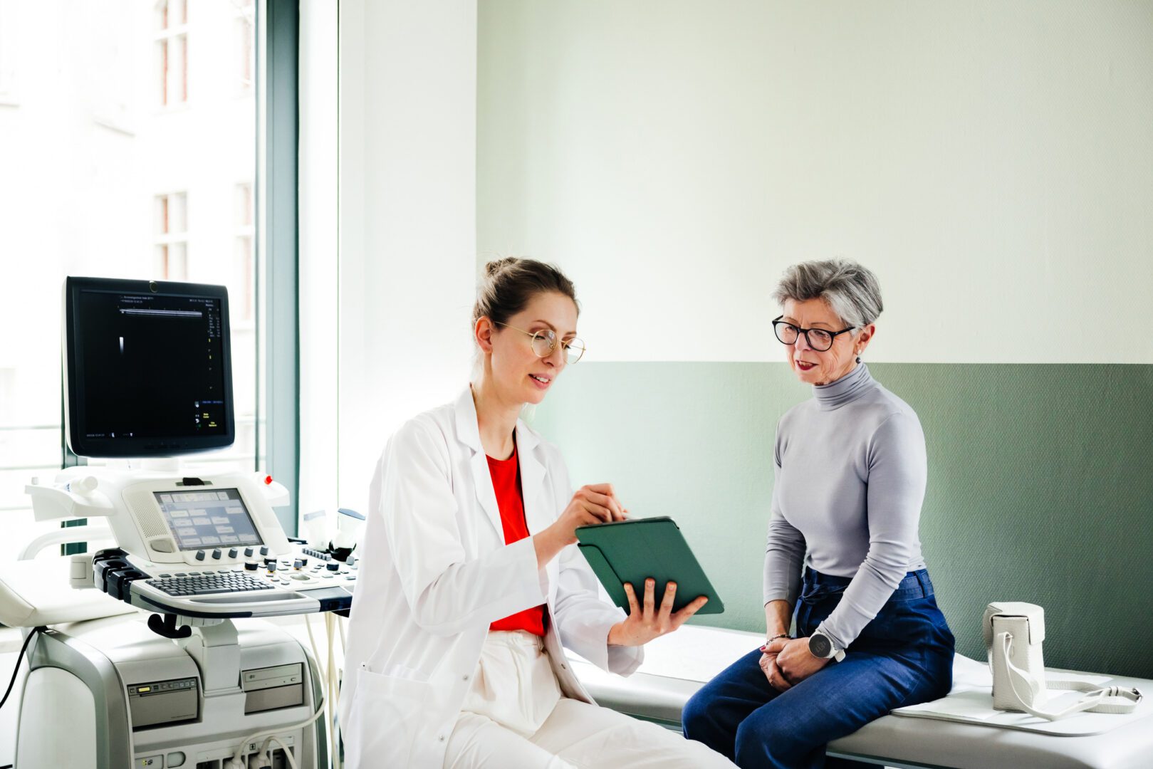 Young female doctor is showing health informations on a digital tablet to an elderly patient. Both are engaged in a discussion while sitting in a medical examination room.