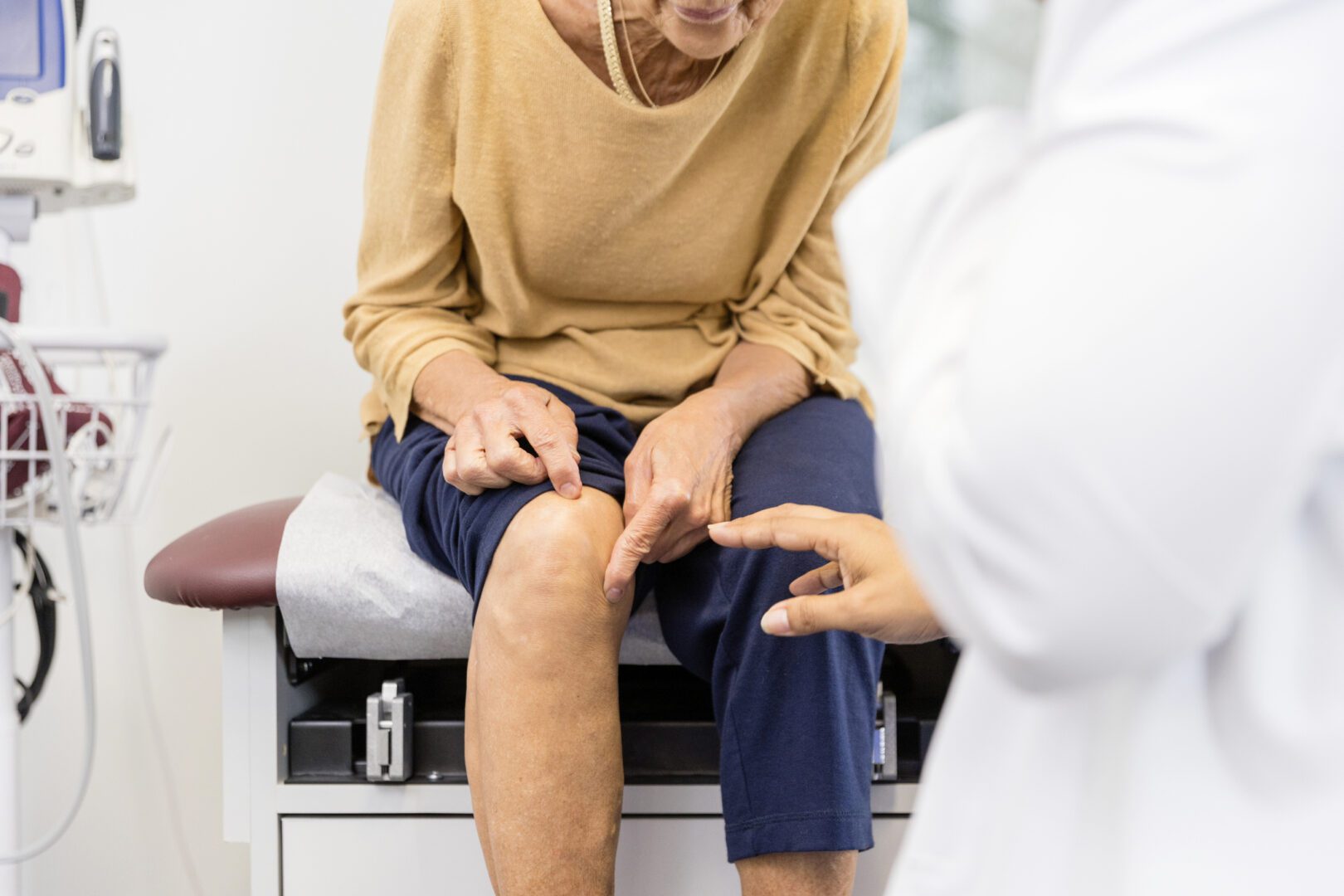 The unrecognizable senior adult woman, sitting on the examination table, shows the unrecognizable female doctor the exact place where her knee hurts.