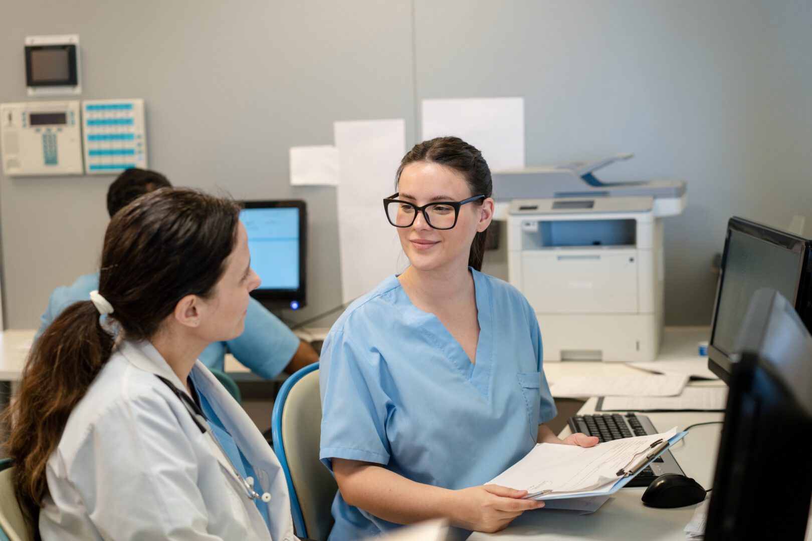 Female admin worker showing a report on clipboard and talking with a doctor working behind counter at hospital. Doctor and administrative worker reviewing a report on a clipboard at the hospital counter.