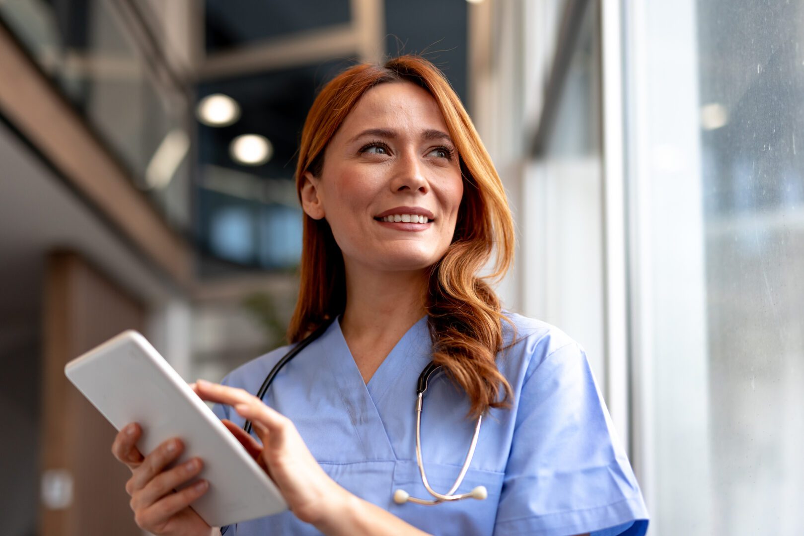 Confident healthcare professional in a uniform holding a digital tablet, looking inspired. Depicts a positive, professional, and tech-savvy medical worker standing by a window in a modern healthcare setting.