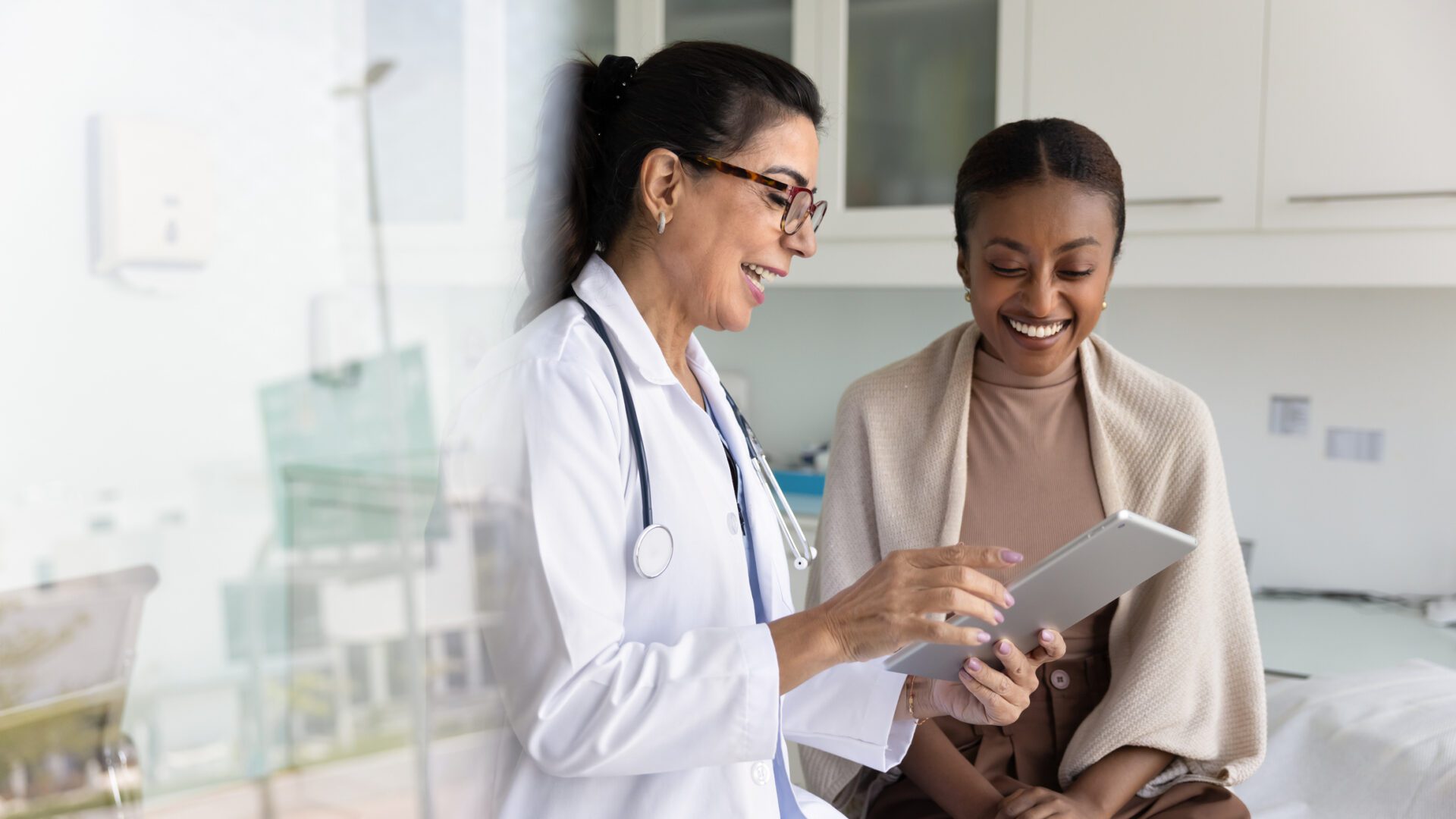 Happy senior practitioner woman and cheerful young African patient woman watching examination medical report on tablet computer, using digital technology, laughing
