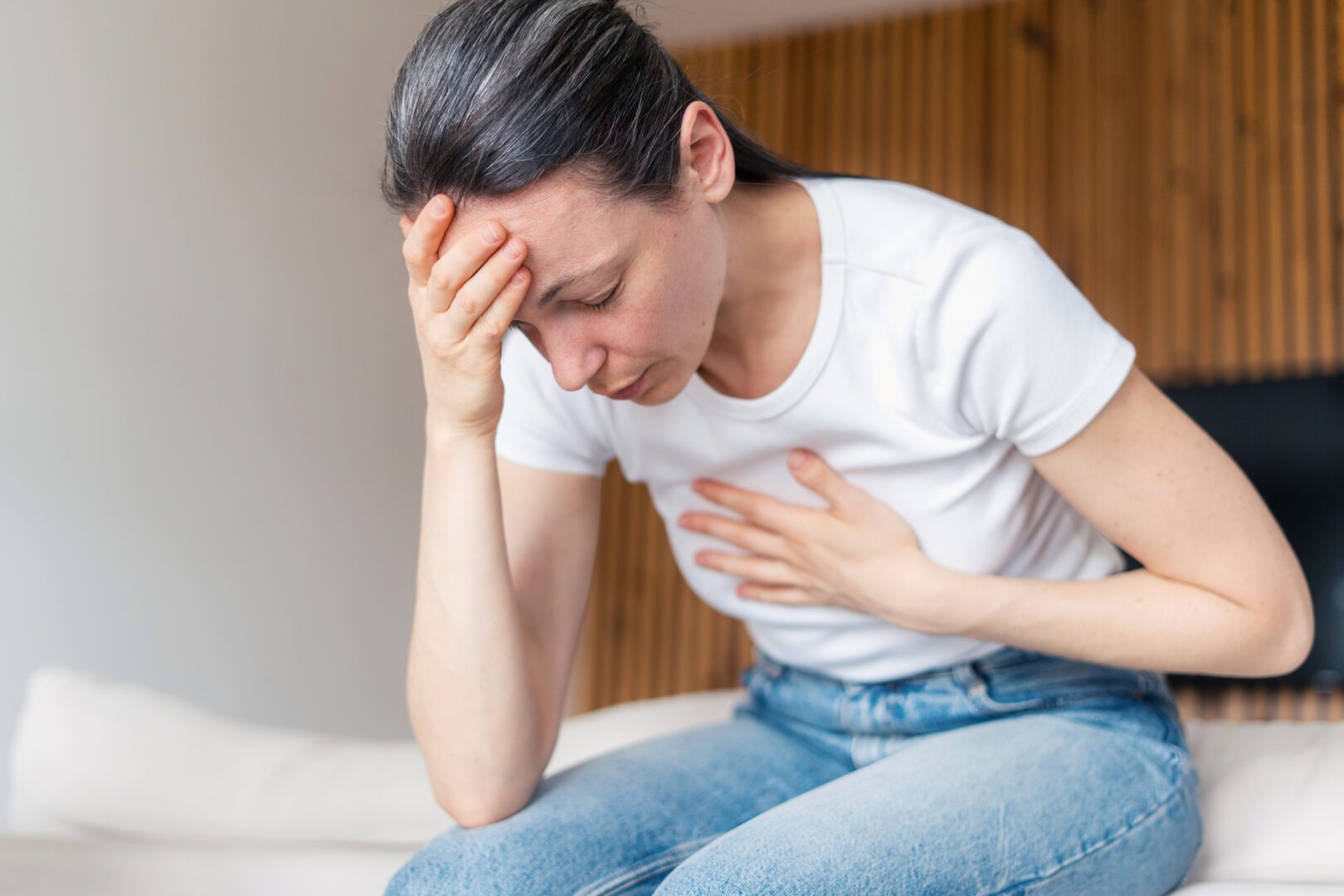 Woman asthma, having heart or panic attack, feeling difficulties with breathe. Female in severe pain and stress, sitting on sofa with closed eyes at home and holding hands on chest and forehead