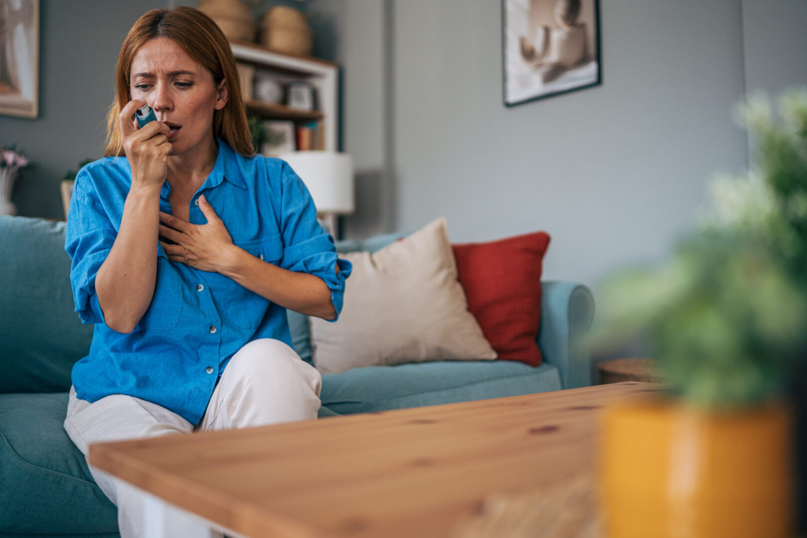 Mid adult woman sitting on a sofa in her living room, using an inhaler to manage asthma symptoms, holding her chest with one hand, reflecting a moment of concern and discomfort
