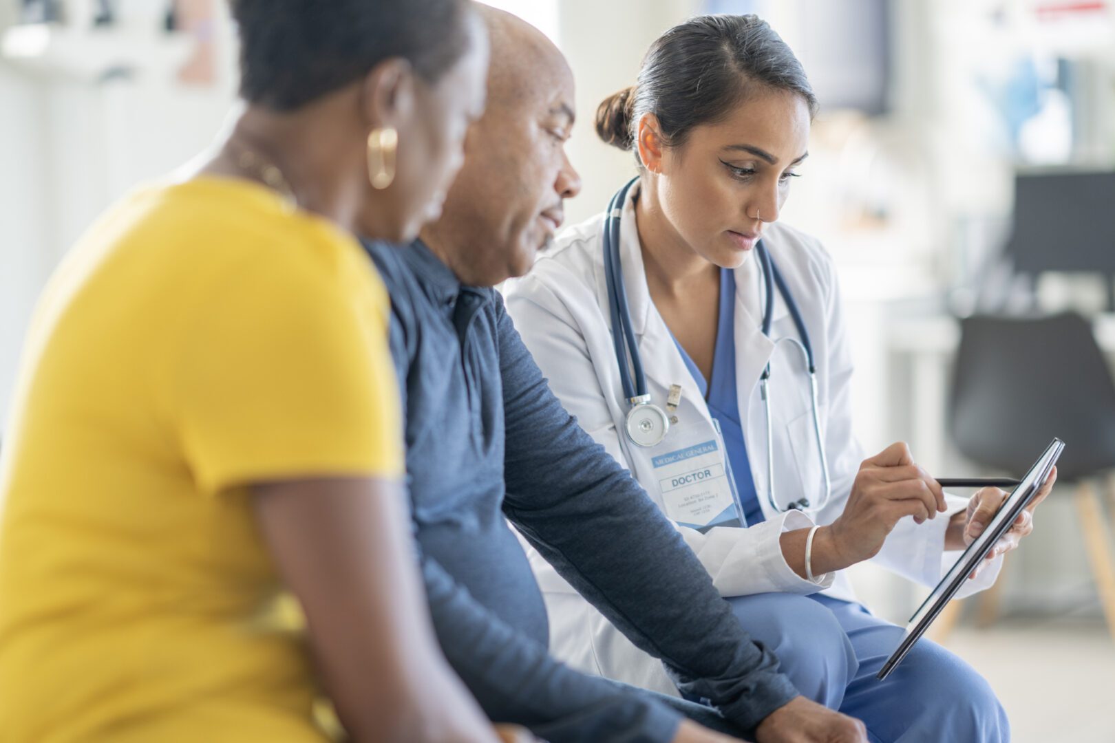 A diverse medical team with a female Asian doctor consults two patients in a bright clinic, using a tablet to review information and discuss care.