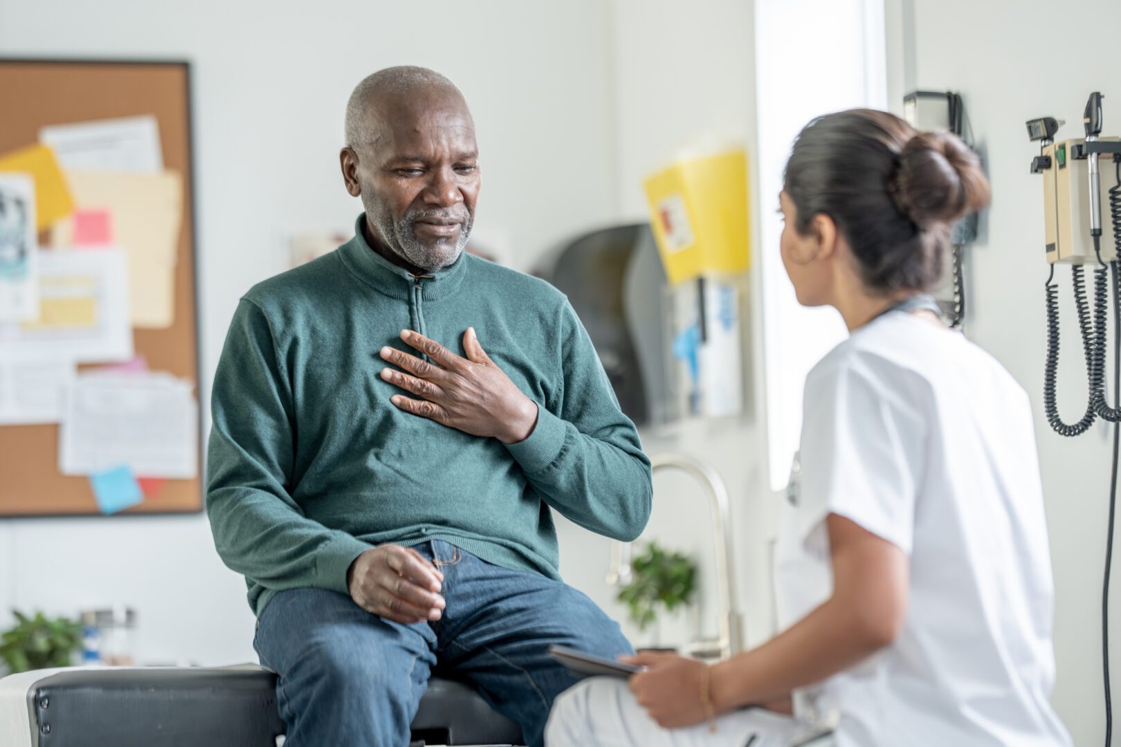 An elderly Black man sits on an exam table, hand on his chest, speaking with a compassionate female nurse in a clinic. A calm, trusting moment highlighting patient care and empathy.