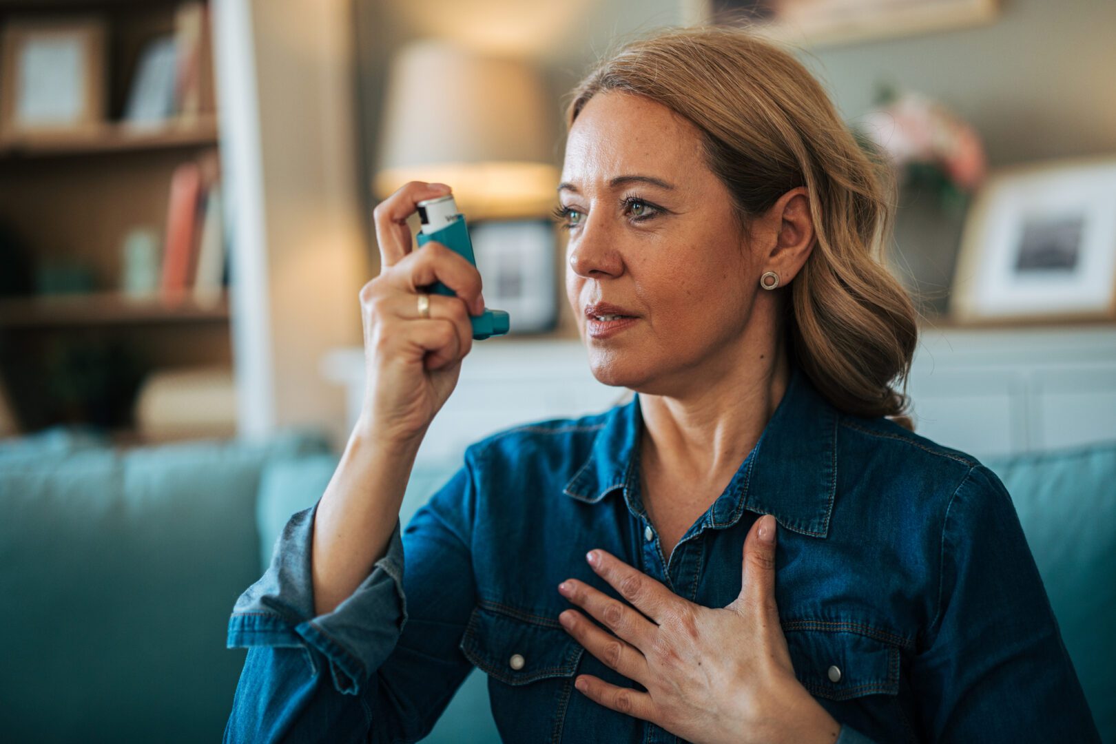 Woman feeling chest pain and breathing difficulty, using a reliever inhaler managing asthma or other respiratory disease symptoms inside her home living room