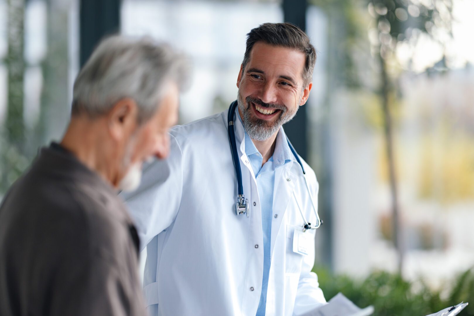 A smiling doctor in a white coat chats with an elderly man in a bright clinic, discussing care plans while holding documents. The scene conveys trust, health, and compassionate medical support.