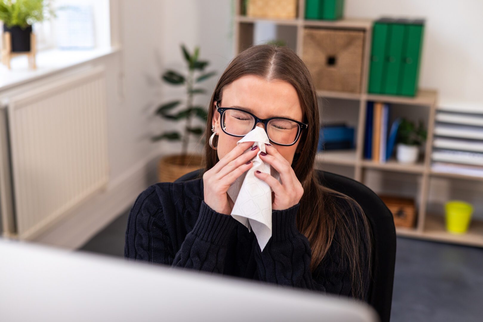 A high-angle view waist-up shot of a young woman with dual sensory loss, wearing glasses, sitting in front of a modern computer, which is out of focus.  She is sneezing into a tissue in a bright and open workspace. Located in Newcastle Upon Tyne.

Videos of this scenario are available