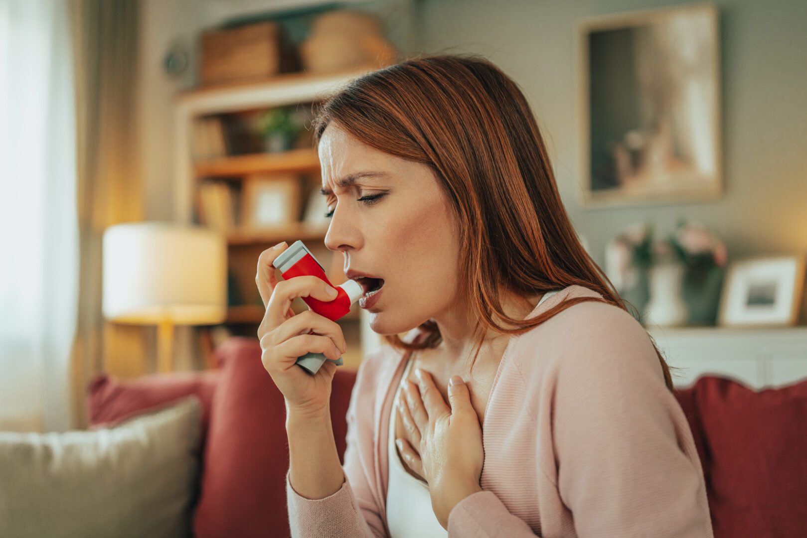 Woman using a red inhaler while clutching her chest on a living room couch, appearing short of breath and concerned. Indoor home setting with soft lighting and blurred background elements suggests a sudden asthma attack or respiratory distress and highlights personal health, emergency response, and self-care.