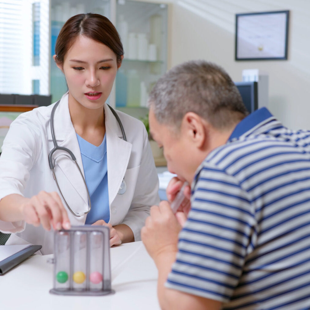 attractive young brunette ponytail female asian doctor wear white coat and stethoscope use triflow training equiptment to improve vital capacity of gray hair old sick man at clinic  - a lung organ model on table