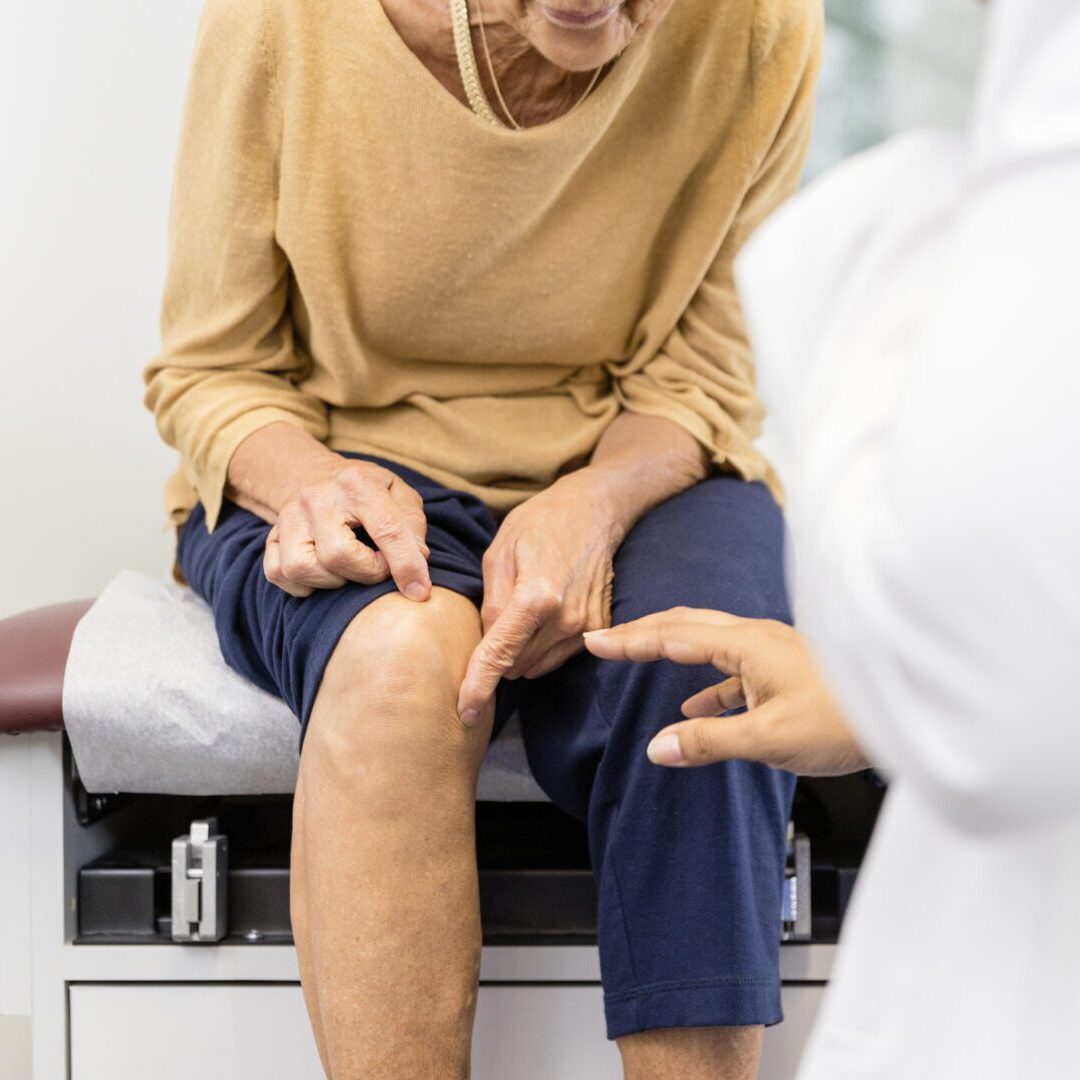The unrecognizable senior adult woman, sitting on the examination table, shows the unrecognizable female doctor the exact place where her knee hurts.
