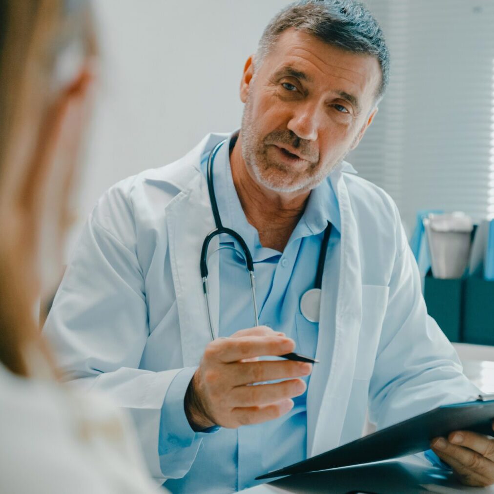 Senior doctor in lab coat using digital tablet discusses medical test results with a patient in clinical office. Medical health care concept.