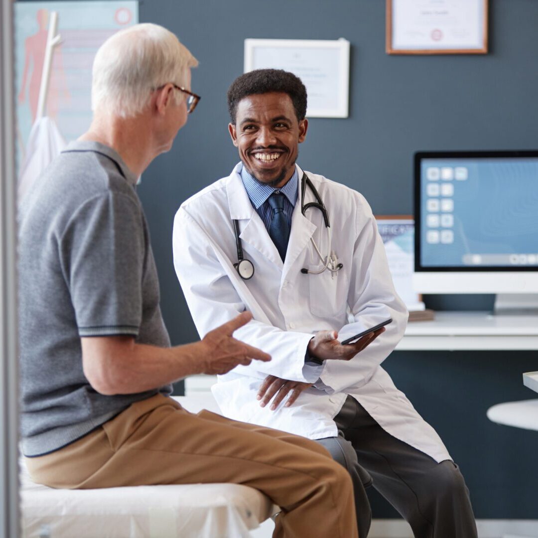 Waist up portrait of smiling African American doctor wearing lab coat and holding tablet while talking to senior patient during consultation in clinic, copy space